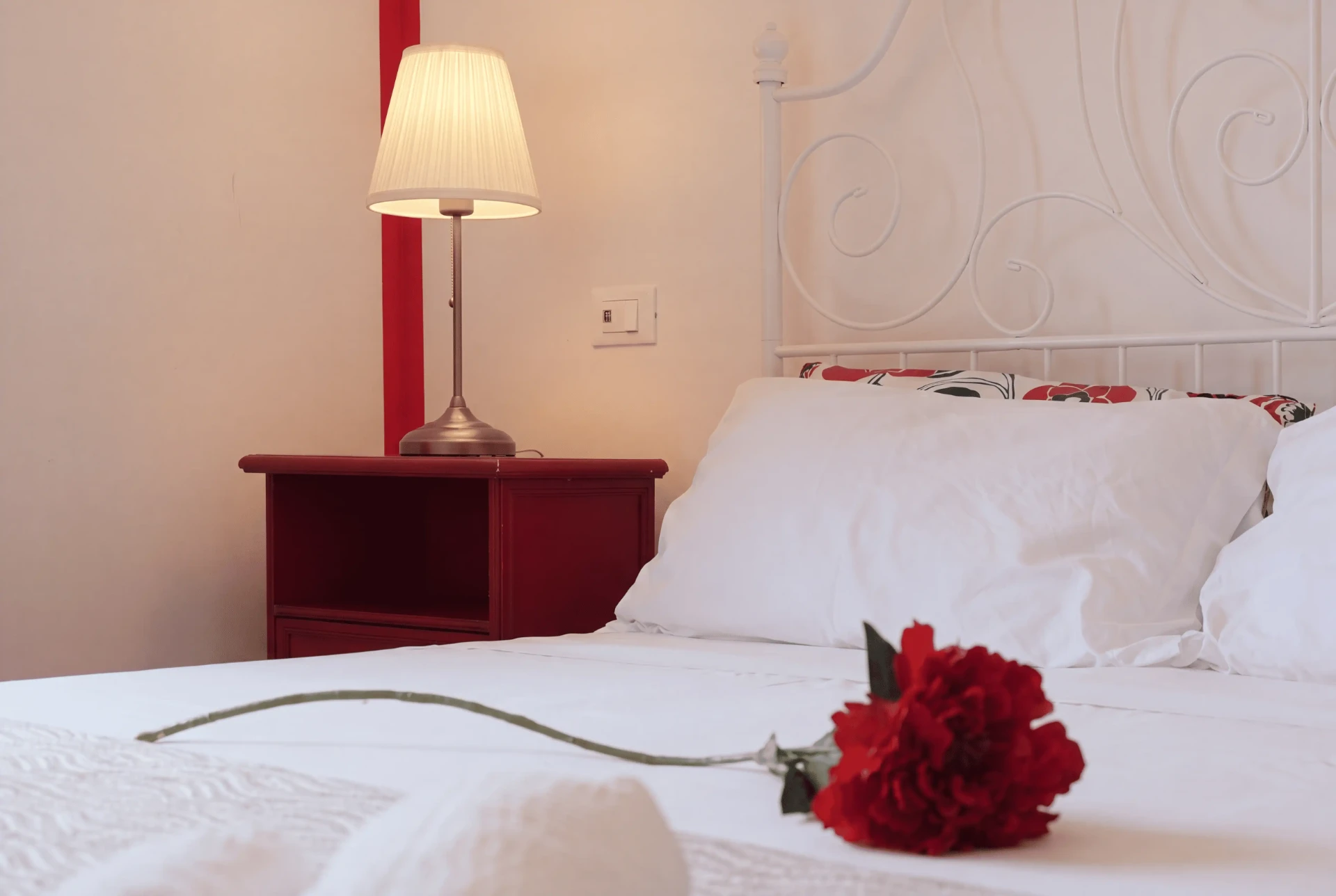 Una rosa rossa sulle lenzuola bianche, con la lampada accesa sul comodino