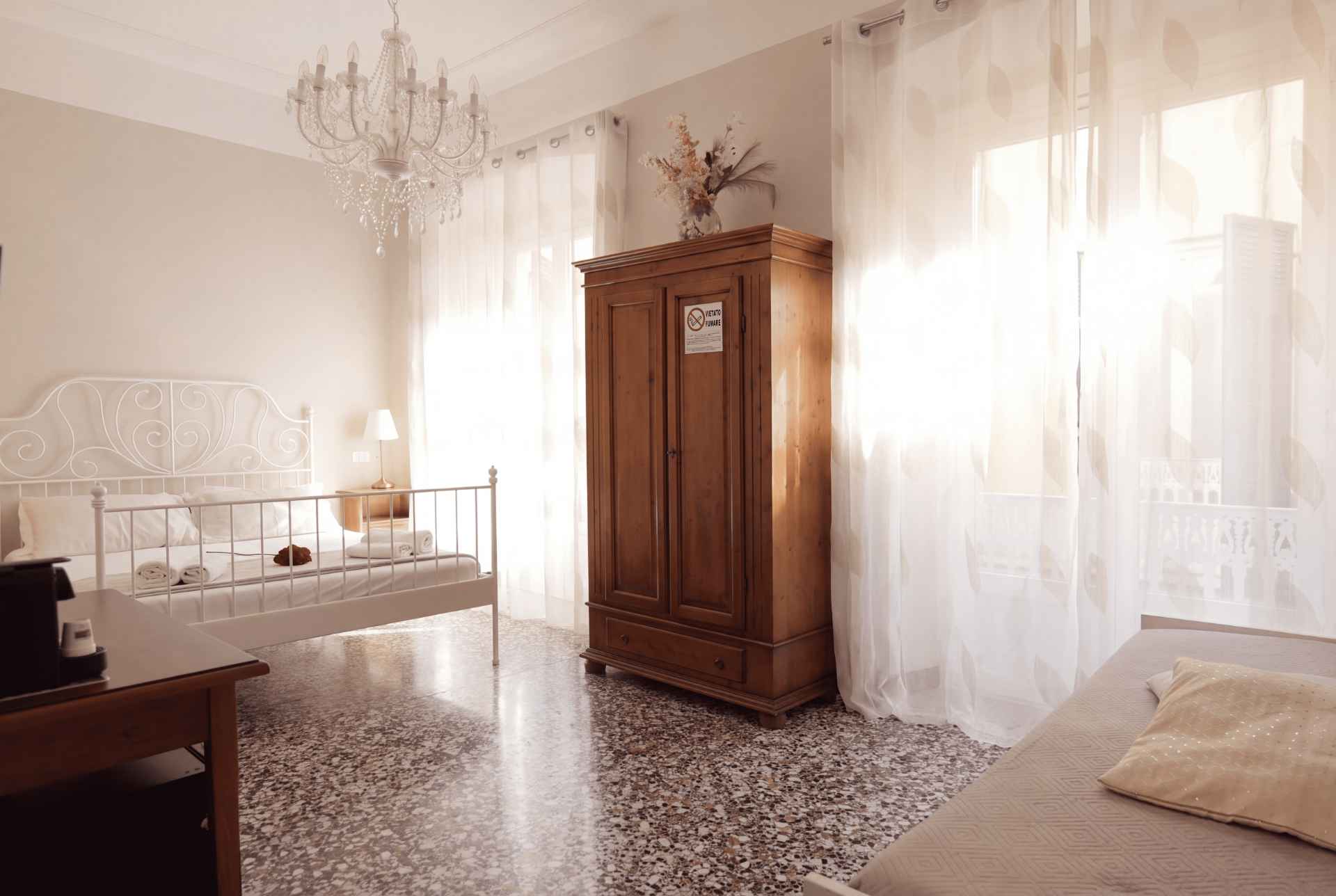 Camera Cachemire: letto matrimoniale in ferro battuto bianco, armadio in legno, tende chiare e pavimento in graniglia sotto un lampadario