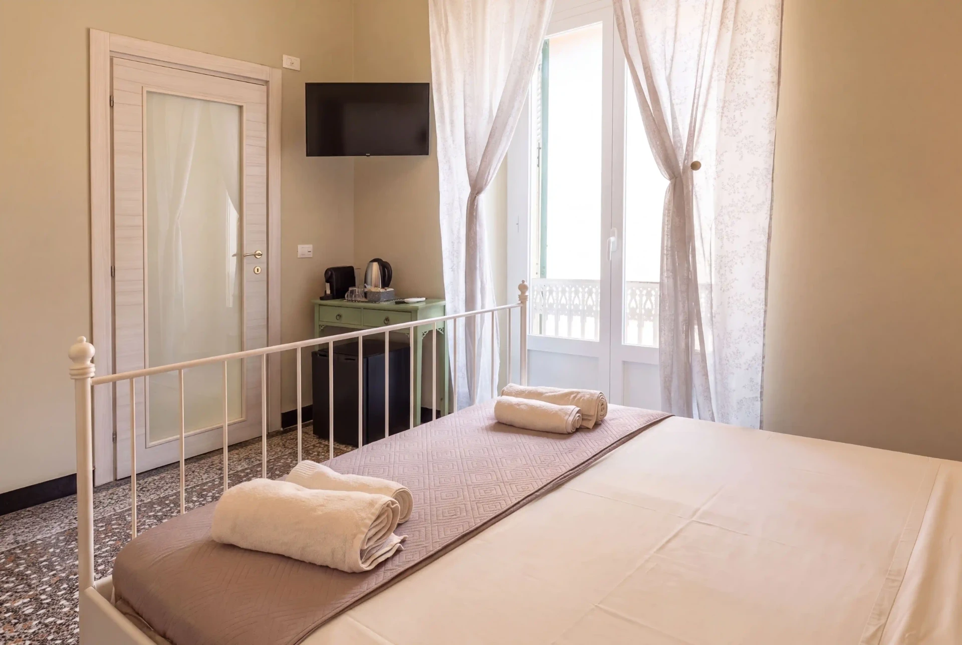 Camera Cannella: letto matrimoniale in ferro battuto bianco con asciugamani arrotolati, portafinestra con tende leggere, scrittoio verde e pavimento in graniglia