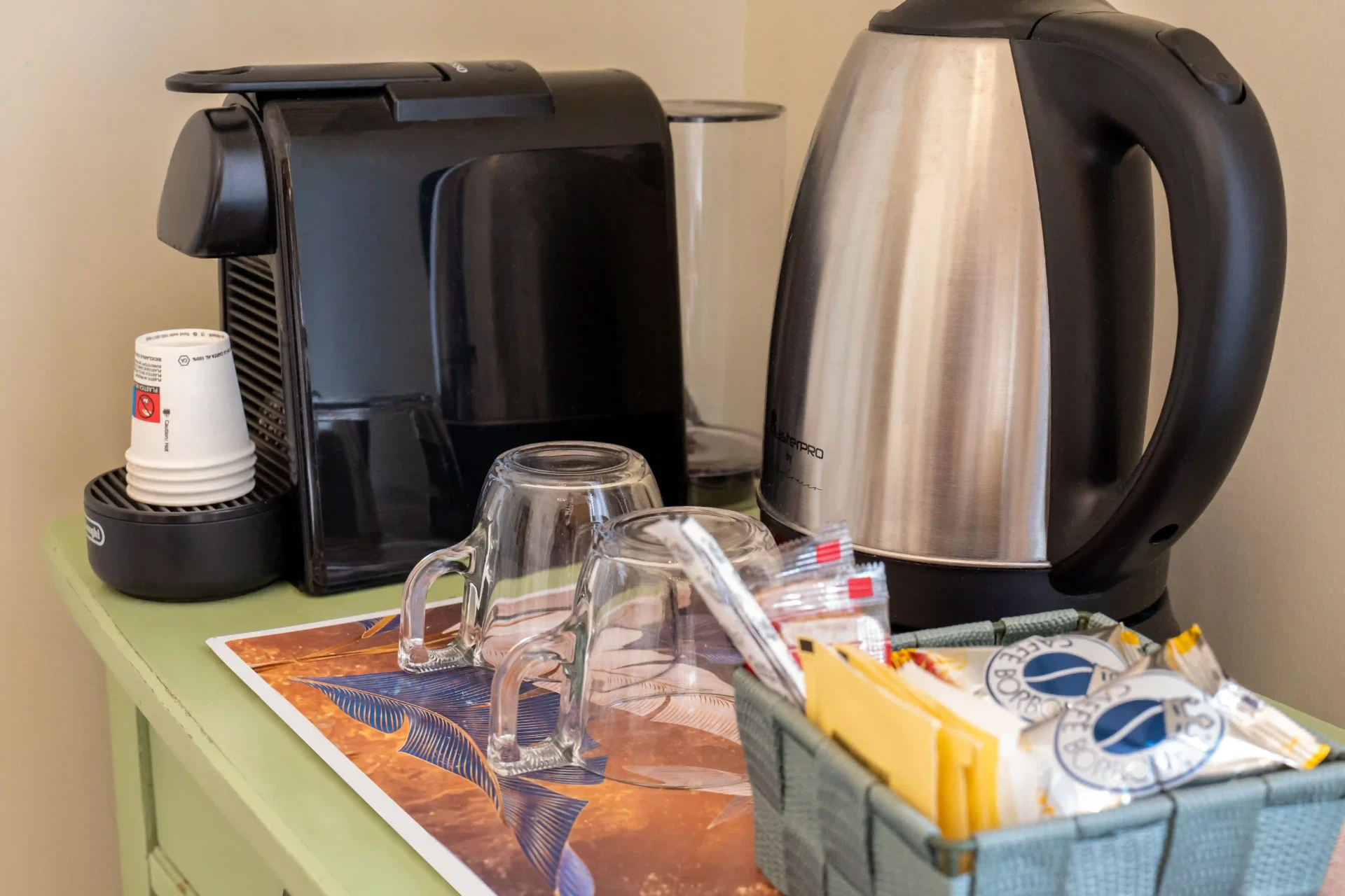 Angolo caffè su un mobile verde: macchina a capsule, bollitore, tazze in vetro e cestino con zucchero e caffè