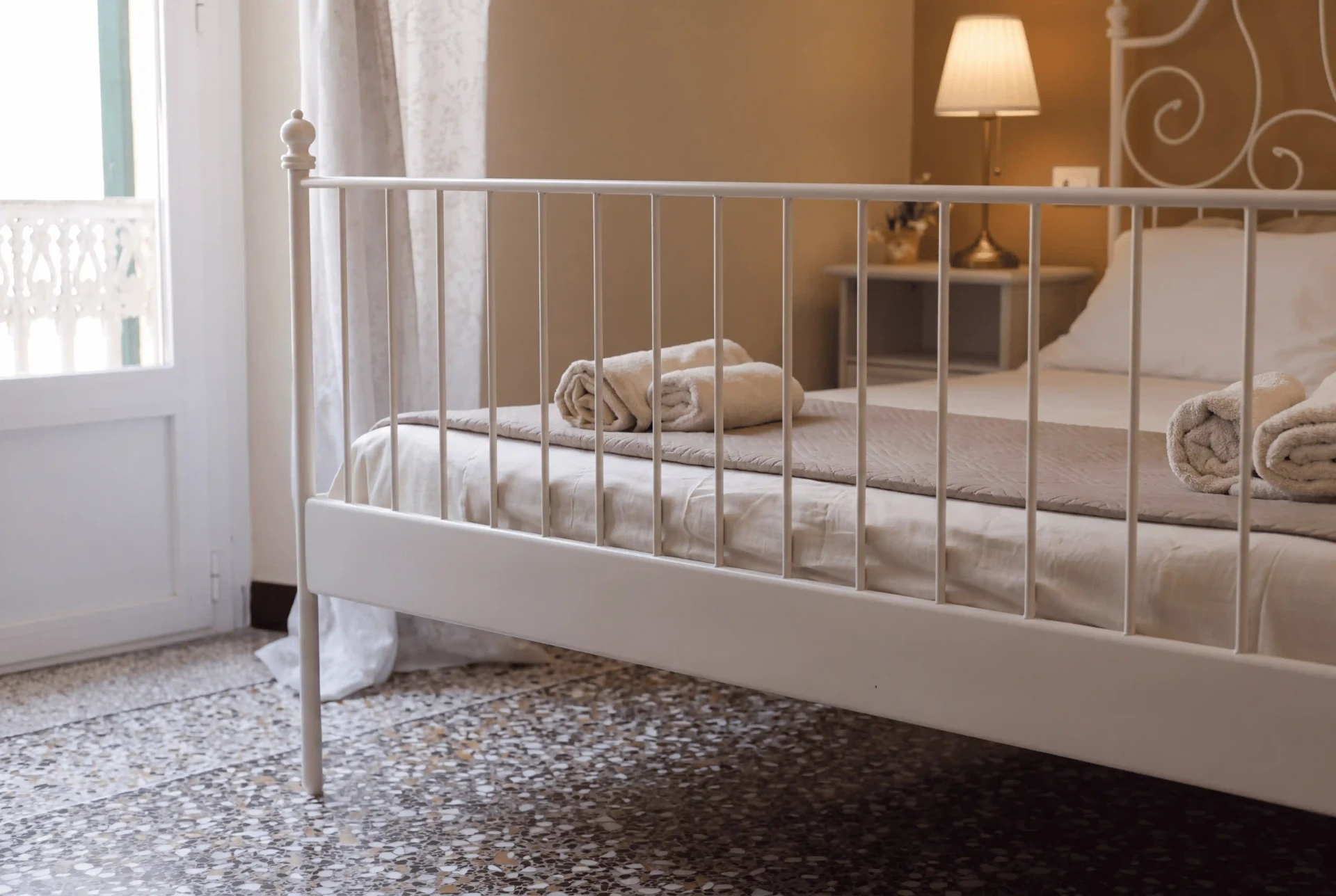 Il piede del letto con la portafinestra, le tende leggere e la ringhiera decorata del balcone