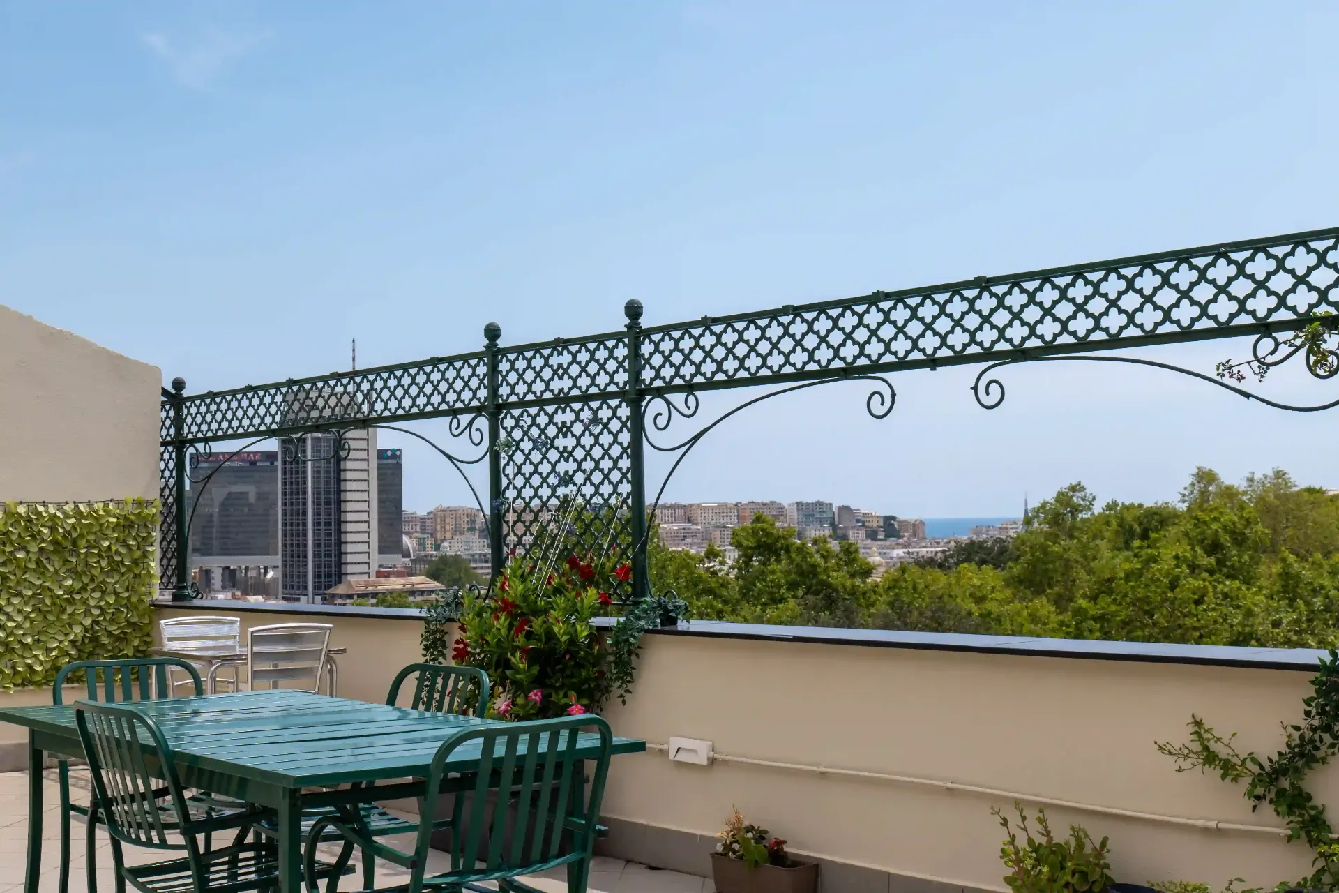 Tavolo e sedie verdi sul terrazzino, piante fiorite e vista sugli alberi e sui palazzi di Genova