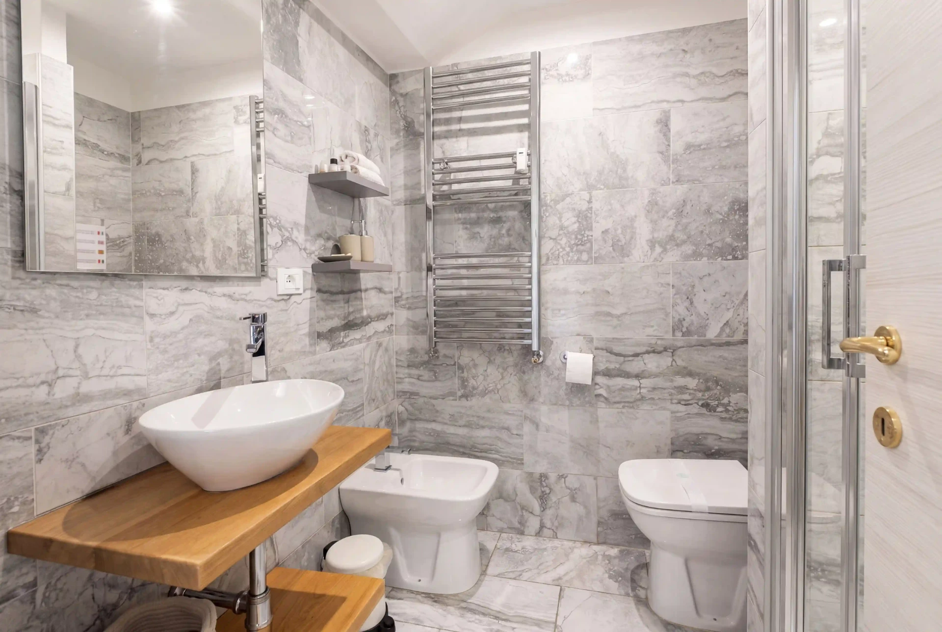 Bagno privato di Oltremare: lavabo d’appoggio su piano in rovere, bidet, WC, scalda-salviette e rivestimento in marmo grigio
