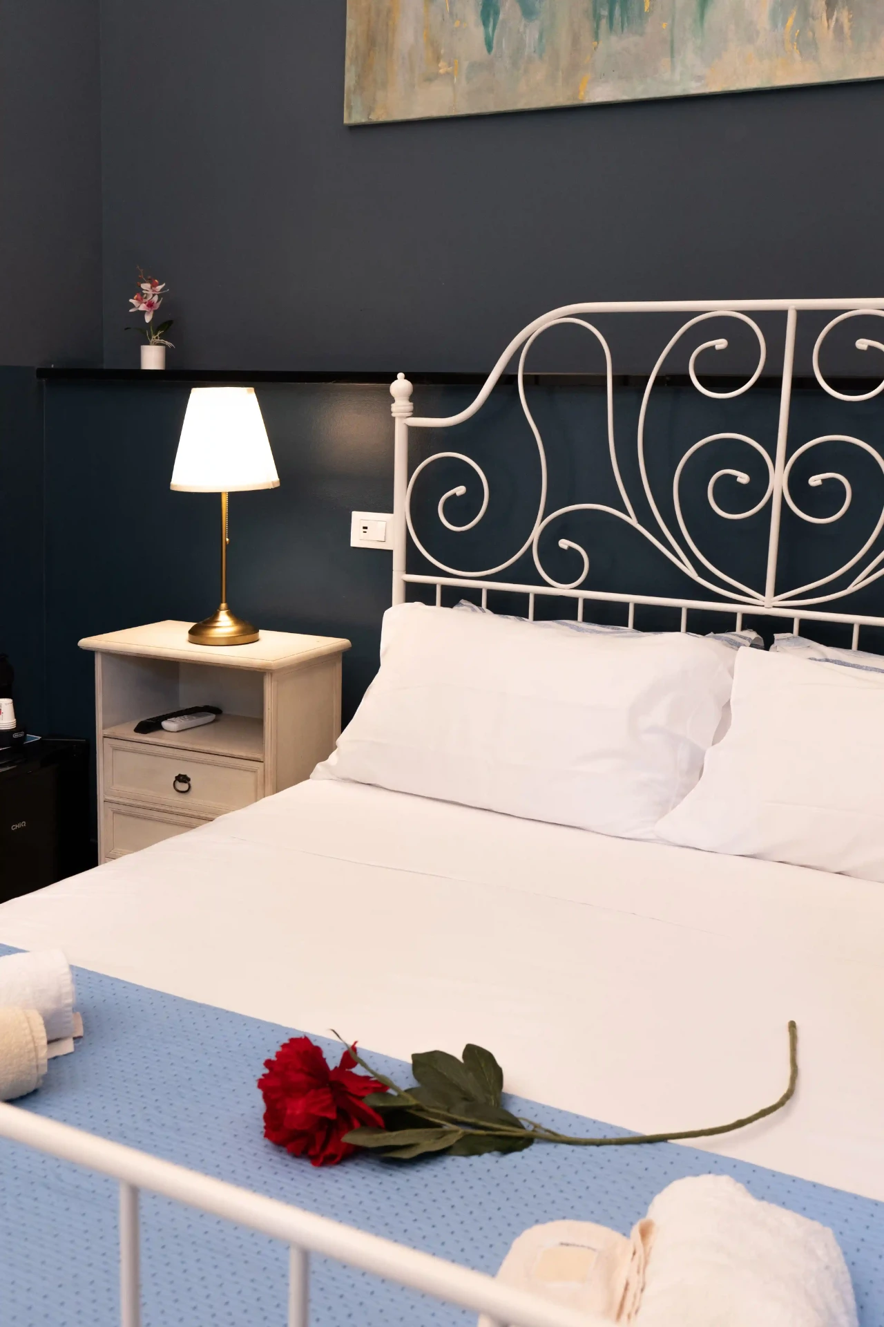 Testiera in ferro battuto bianco, comodino con lampada e rosa rossa sul letto