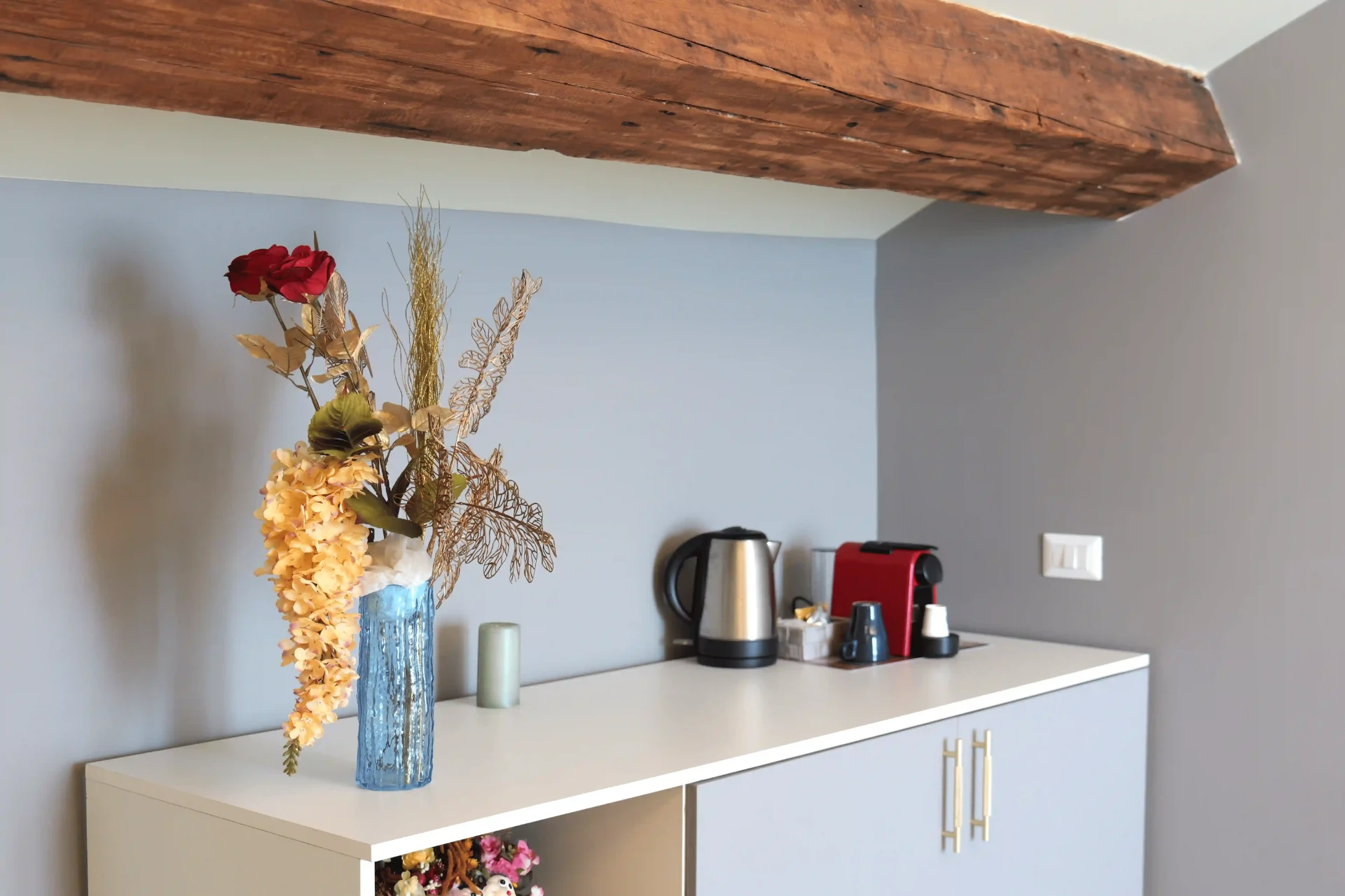 Piano bianco con bollitore, macchina a capsule rossa e fiori secchi, sotto una trave in legno