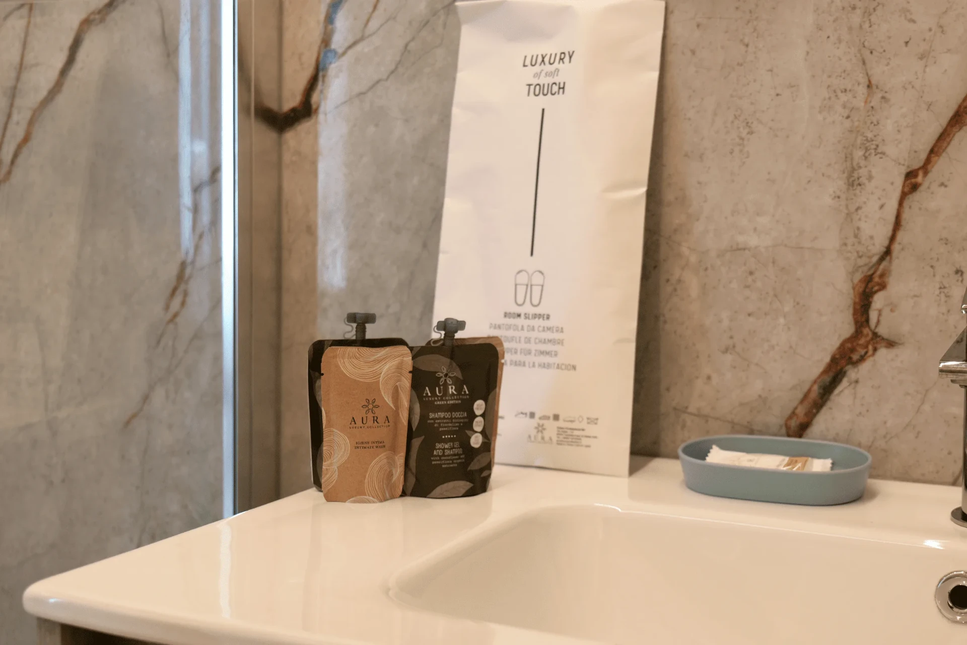 Lavabo bianco con prodotti di cortesia, ciabattine e rivestimento in marmo venato