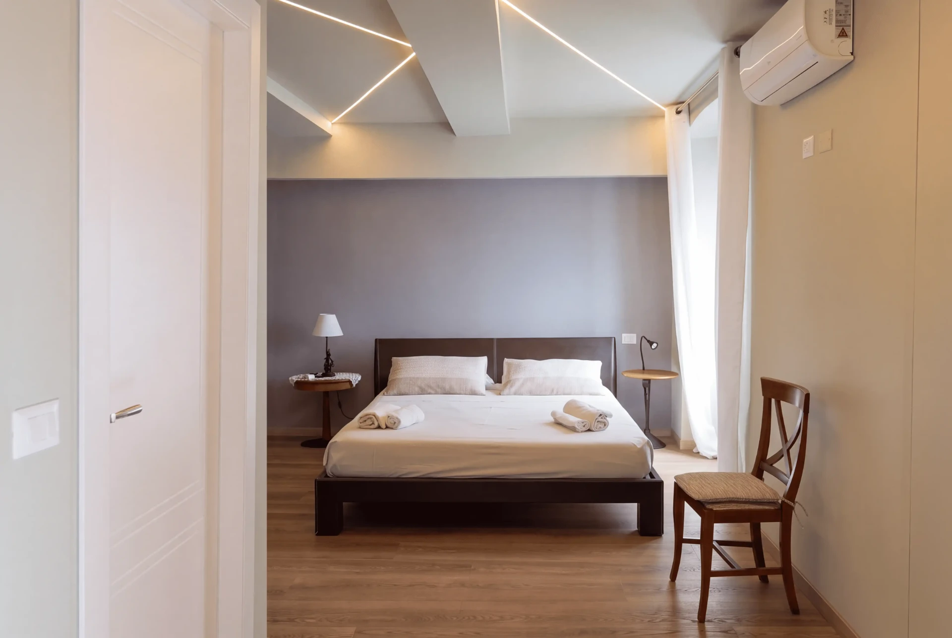 Suite in mansarda: letto matrimoniale basso, sedia in legno, portafinestra con tende bianche e luce a taglio sul soffitto